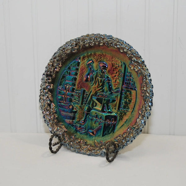 A front view of the Fenton American craftsman carnival glass plate number 3. Circa 1972. It features James Read who is a blacksmith and working in his shop.