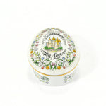 Shown is a oval or egg shaped music box produced by Franklin Porcelain or Franklin Mint. On the top/cover there are delicate flowers and leaves around the edge with the song title 'Somewhere My Love', written in cursive around a castle which is in the center. The edge of the top is painted in gold. It is on a white background. The body and top are also white. Part of the floral decoration can be seen on the side of the bottom of the music box.
