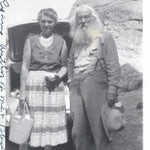 Shown is a vintage  black and white photo of an unknown woman who is dress in a  skirt and sleeveless top and holding a handbag in her right hand. On her left is author and prospector Ed Ryan. He has light colored hair that is shoulder length and a full beard and mustache. He is wearing a long sleeve shirt, long pants with suspenders. He is holding a wide brim hat in his left hand. On the left margin of the photo, written in ink it states Ed Ryan author of Me and Black Hills.