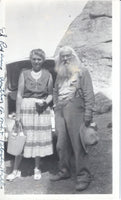 A full view of the vintage black and white photograph of an unknown woman and author Ed Ryan. They are standing in front of a car that has the trunk open and a very large rock out cropping.