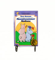 Shown is the front cover of the Moominsummer Madness paperback book. There are two hippos on the cover and the one on the right is holding a bouquet of flowers. It is propped up on a black metal stand.