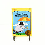 Shown is the cover of the paperback book Tales From Moominvalley. There is a disgruntled looking hippo in a black top hat, sitting on a chair at a outside table and they are holding a drink. There is a yellow and orange umbrella shading him. The book is propped up on a black metal stand.
