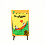 Shown is the cover of the paperback book, Moominpappa at sea. There are two hippos in an orange row boat. The one on the left is wearing a hat and holding a fishing net. They are heading up a large wave.