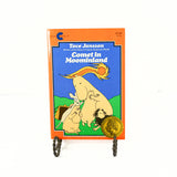 Shown is the front cover of the paperback book Comet in Moominland by Tove Jansson. There is an adult hippo who is comforting what appears to be a kangaroo and a small hippo. There is also another small hippo with their hands over their eyes and what appears to be a small dog like creature. The book is propped up on a black metal stand. 