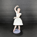 Shown is a Royal Copenhagen B & G (Bing & Grondahl) porcelain figurine of a ballerina. She has one arm raised up and her left arm is partially across her waist. She is dressed in a knee length white dress the has delicate flowers on it. She also has flowers in her hair. She is wearing light brown ballet slippers. The round base is a delicate gray color.