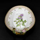 Shown is a Royal Copenhagen oval dish. It made of white porcelain and is hand painted with a sprig of oregano. Around the edge of the dish there is also hand painted florals and design. The outer edge of the dish is slightly serrated and edged in gold paint. There is one handle which is also porcelain and made to look like a rounded trig. There are two applied porcelain flowers on the right side of the handle. The dish is on a black background. 