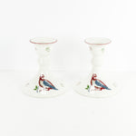 Shown are a pair of vintage Johnson Brothers 12 days of Christmas candlesticks. These feature a pair of blue and red partridges on each candlestick along with some green and red pears. The candlestick are white and shown on a white background.