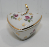 The photo shows the lidded ceramic dish. It has roses with leaves and gold decor on the lid and bottom part. The lid also has a delicate handle that is white and trimmed in gold. The lid is also trimmed in gold.
