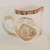 Shown is a close up of the Old English Staffordshire Ware small pitcher celebrating the Lincoln Sesquicentennial. On the front it shows a log cabin which was Lincoln's birthplace in Kentucky. The creamer is a creamy white color with a brown or sepia tone transferware pattern.