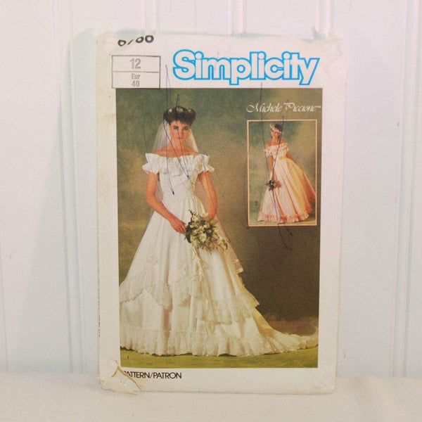 Shown is the front of the paper envelope for vintage Simplicity 6766 sewing pattern, c. 1984. There is a color photo of a young woman in an example of this wedding dress, which is off the shoulder and short sleeved. There is an inset color photo of another young woman wearing a different version of the dress in a pale pink color. The pattern is designed by Michele Piccione. There are some ink scribbles on the front.