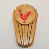 Shown is a vintage wood knife holder. There is a round wood disk on the upper part where there is a painted red chicken with small yellow hearts on the body. It is shown on a white background.
