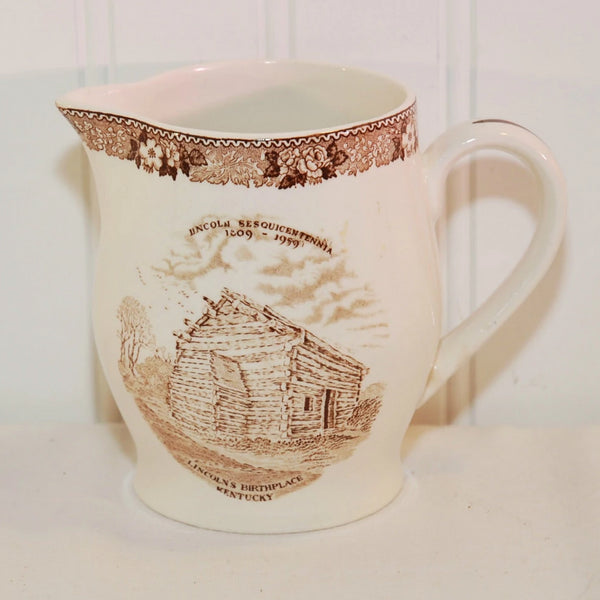 Shown is the vintage Nancy Lincoln Inn commemorative creamer, c. 1959. It is a creamy white creamer with brown transferware decoration. The top border has a floral motif. In the center it states Lincoln Sesquicentennial, 1809-1959. Below that it has an illustration of Abraham Lincoln's birthplace log cabin. Below that it says Lincoln's Birthplace, Kentucky. The spout is on the left and the handle on the right.