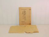 In this photo the paper instruction sheet is seen propped up on a white wall with the tissue paper sewing pattern laying in front of it. It is also on a white background.