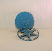 Shown is a metal movie reel in front of a blue metal movie reel canister. Both pieces were made by Compco Corporation.