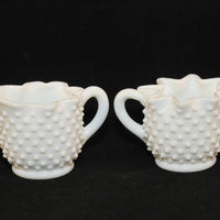 PIctured are a vintage Fenton art glass creamer and sugar set from circa 1967. They are white in color with a hobnail pattern and a star crimped edging. Each have milk glass handles.