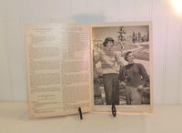 Another photo of the vintage knitting book interior. Once again on the left are instructions for knitting. On the right is a black and white photo of two young women in long sleeve knitted sweaters. Behind them is a snowy scene with snow covered pine trees.
