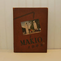 A front view of the hardback circa 1942 Ohio State University Makio yearbook. The cover is brown in color and the print in black. There is a color photo on the front, possibly of the entrance to the campus. The photo show two large square pillars that are quite tall and each has a large ball shape feature on top. There is an 1940's vehicle to the left of the left pillar and an assortment of students in front and in between the two pillars.