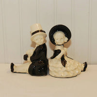 A pair of vintage bookends featuring a Victorian style boy and girl sitting with their backs to each other. The boy is wearing a white tall hat with a black band, a black waistcoat and black shoes. The girl is wearing a black hat that ties with a black ribbon under her chin. She is wearing a black shawl and shoes, her ruffled dress is white.