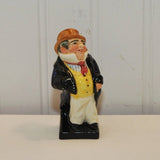 The photo shows the vintage Royal Doulton bone china figurine, Captain Cuttle. Captain Cuttle is quite dapper, he is wearing a brown top hat. His shirt has a high white collar that hides his neck with a space for his chin, the rest of the shirt is blue and white vertical stripes. He has a red tie on. He has a black overcoat on that is open. He also has a yellow vest with black button on over his shirt. He is wearing baggy white pants and black shoes. He is standing on a black platform.