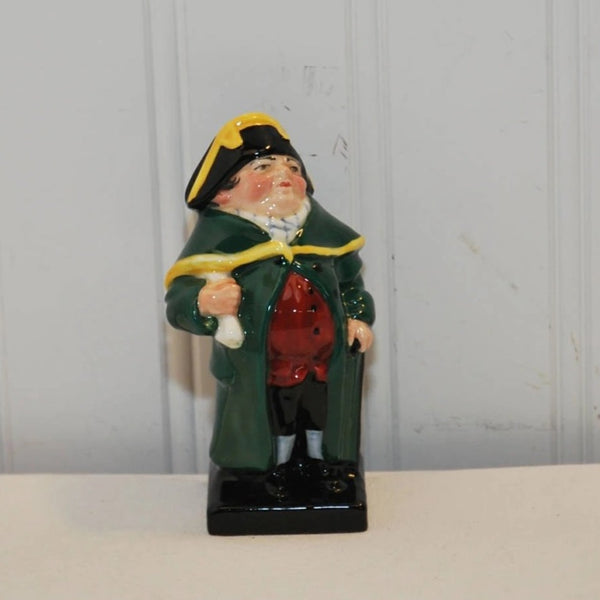 The photo shows a vintage Royal Doulton bone china figurine Bumble. He is wearing a black hat with yellow trim. His overcoat is green with yellow trim. He is holding a white object in his right hand. His collar is white and he is wearing a red vest with black buttons. He has black knickers and shoes on. He has white socks on. He is standing on a black platform.