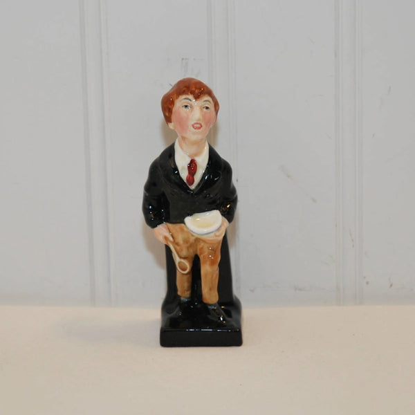 A front view of the vintage Oliver Twist figurine that was made by the Royal Doulton company. It is made of bone china and dates between c. 1922-1983. The figurine depicts a boy in a black suit coat, white shirt with red tie and brown pants. He has black shoes on. He is standing holding a white bowl in his left hand and a wooden spoon in his right.