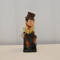 The photo show the vintage Royal Doulton Sam Weller Figurine c. 1939. The figurine is hand painted and is shown sitting, wearing a brown top hat slightly tipped on his head. He has black hair. He has a red kerchief tied around his neck and is wearing a white shirt that has a yellow and brown stripped button vest. He has brown pants, reddish brown pant protectors on his calves and black shoes.