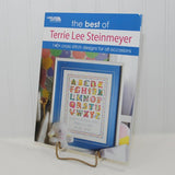 This photo shows the front cover of the cross stitch paperback, The Best of Terrie Lee Steinmeyer. 140+ cross stitch designs for all occasions. It was published by Leisure Arts. The book is propped upright on a small stand.