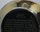 A close up of the Fenton paper label the shows a glass blower  and states Authentic Fenton Handmade in a black oval in front of the craftsman. The label is gold and black.