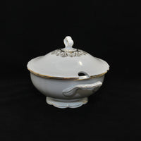 This photo show a side view of the soup tureen. There is a half circle cut out on the lid for a soup ladle. There is a delicate gold decor around the handle of the lid. There is gold trip around the edge of the lid, the top rim of the tureen, the outer edge of the handles and the base of tureen. 