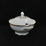 This photo show a side view of the soup tureen. There is a half circle cut out on the lid for a soup ladle. There is a delicate gold decor around the handle of the lid. There is gold trip around the edge of the lid, the top rim of the tureen, the outer edge of the handles and the base of tureen. 