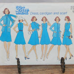 A close up of the front of the envelope for vintage Simplicity 6850 is shown in this photo. It is resting on a small metal stand. There six illustrated women, all dress in sky blue outfits that can be made with this pattern. The pattern states "Go Every-Where, Dress, cardigan and scarf".