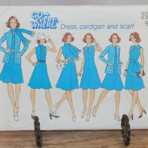 A close up of the front of the envelope for vintage Simplicity 6850 is shown in this photo. It is resting on a small metal stand. There six illustrated women, all dress in sky blue outfits that can be made with this pattern. The pattern states "Go Every-Where, Dress, cardigan and scarf".
